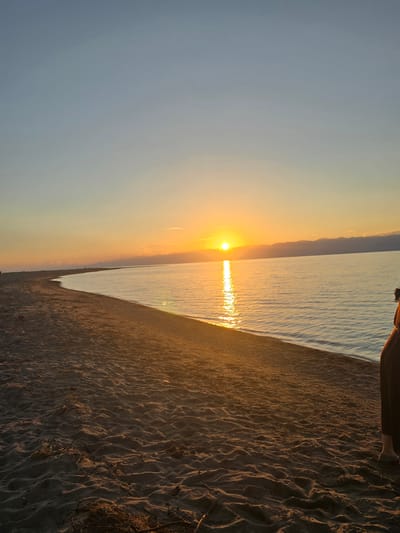 This is a vertical, candid, eye-level photograph taken at sunset on a sandy beach, likely at Boz Beshik, Kyrgyzstan. The sun, a bright orb of light, is low on the horizon, casting a golden glow across the scene. Its reflection shimmers on the calm, light blue water of a large body of water, possibly a lake or sea, that stretches towards the horizon.
The foreground is dominated by a sandy beach, marked with undulating patterns created by wind or waves. The sand is a warm, earthy brown, illuminated by the setting sun, which creates long shadows. The sand appears dry and textured, with a few dry twigs or grass scattered across it.
In the upper portion of the image, a person's legs and feet are visible, wearing dark clothing. They are seated or kneeling on the sand, looking out towards the sunset. The person's presence suggests a moment of quiet contemplation or enjoyment of the natural scenery.
The sky transitions from a clear, pale blue at the top to a soft orange and yellow hue near the horizon, a typical characteristic of a sunset. In the distance, a hazy, dark silhouette of hills or mountains can be seen against the brightly lit sky. The overall mood of the image is peaceful and serene, capturing the beauty of a natural landscape at the end of the day. The time of day is clearly sunset, and the weather appears clear and calm. There is no visible text in the image.