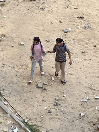 Two girls are standing on a sandy surface littered with debris, possibly in Khan Yunis. The girl on the left wears a striped shirt and beige pants and is barefoot. The girl on the right wears a pink shirt and grey pants with pink sandals. A gray railing with debris and plants are visible in the bottom right corner. The environment appears to be sandy with various small debris scattered.