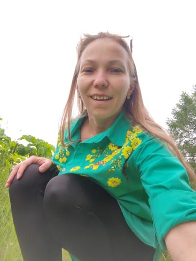 A woman with long blonde hair, wearing a green shirt with yellow floral embroidery and sequins, and black leggings, is crouching outdoors in a grassy area. She is smiling at the camera. Behind her, there are green plants and trees, suggesting a natural or park setting. The sky is overcast. The scene is captured from a low angle, with the woman's face and upper body dominating the foreground. No specific landmarks of Chukhlem, Russia, are visible.
