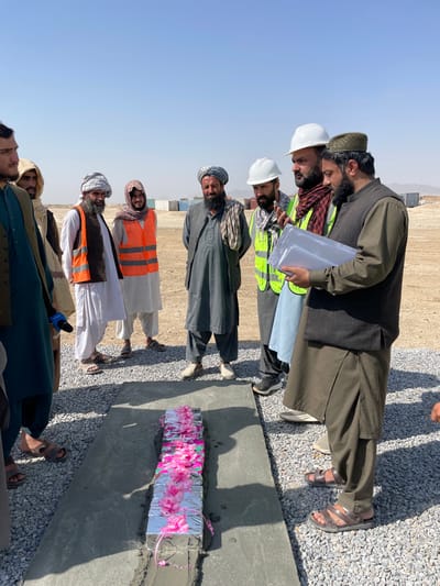 This image depicts an outdoor gathering of nine adult males on a construction site in Pir Dust, Afghanistan. The scene is illuminated by bright daylight under a clear, pale blue sky, with strong shadows indicating direct sunlight.
In the foreground, a rectangular object, approximately 0.6 meters long, wrapped in reflective silver material and adorned with multiple pink fabric flowers and ribbons, is embedded within a freshly poured concrete slab on a ground of light gray gravel. This object appears to be a ceremonial cornerstone or time capsule.
Eight men are positioned around this central object. On the right, a man wearing a dark green shalwar kameez and a patterned cap holds clear plastic folders containing documents and appears to be speaking or explaining. To his left, another man in a white hard hat and a lime green safety vest gestures with his right hand while looking at the documents. Further left, a man in a gray shalwar kameez and a turban observes. Another man, wearing a white hard hat and a dark scarf, stands next to him.
On the left side of the frame, three men are partially visible. One man in a dark teal shalwar kameez holds what appears to be a microphone near his waist. Another man wears a white shalwar kameez and an orange safety vest. All visible individuals are men, many with beards, and some wear traditional Afghan clothing, turbans, or headscarves.
The immediate ground is covered in light gray gravel around the concrete slab. In the mid-ground, behind the individuals, the terrain is dry, dusty, and light brown. Several stacked shipping containers of various colors are visible in the background, suggesting a storage or operational area within a larger construction zone. The distant horizon is flat and arid.
