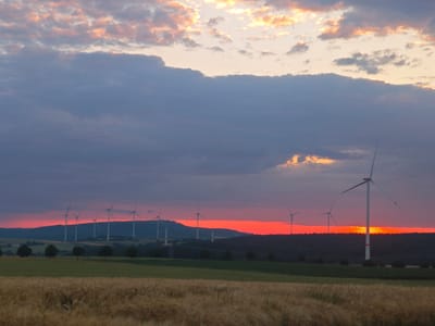 This photograph, taken at sunset in Lügde, Germany, captures a serene rural landscape. The sky is a dramatic display of color, with a band of intense orange and red near the horizon, suggesting the sun is setting behind a distant hill. Above this, the sky transitions to darker blues and purples, with scattered clouds tinged with pink and orange from the fading sunlight.
Dominating the middle ground are several wind turbines, silhouetted against the vibrant sky. They are positioned along a gently rolling hillside, suggesting a wind farm in operation. The turbines vary in size and placement, with one prominent turbine in the foreground on the right side of the image.
In the foreground, a field of golden wheat or barley stretches across the frame, its stalks catching the last light of the day. Behind this, a strip of green field is visible, followed by a line of dark trees and shrubs that form the base of the hills where the wind turbines are situated. The overall impression is one of natural beauty and renewable energy coexisting in a peaceful, rural setting. There are no people or animals visible in the frame.