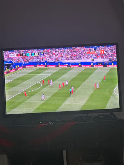 The image shows a television screen displaying a live soccer match. The score is 0-0 between Switzerland (SUI) and Bosnia and Herzegovina (BIH) at 19:57. The game is being played in a stadium with a crowd in the background. There are advertisements for "Lenovo" and "FIFA WORLD CUP RUSSIA" visible on the screen. The television is placed on a dark surface, possibly furniture, in a dimly lit room. The overall scene suggests a casual viewing of a sporting event. There is no indication of the specific location in the city of Mainburg, Germany.