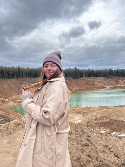 A young woman wearing a beige trench coat and a gray beanie stands in front of a large quarry or canyon filled with turquoise water. The sky is overcast with dark clouds. The quarry walls are made of brown earth and vegetation. Trees line the back of the quarry opposite the woman. The location appears to be a man-made excavation, not a natural feature. It is located in Ivlevo, Russia.