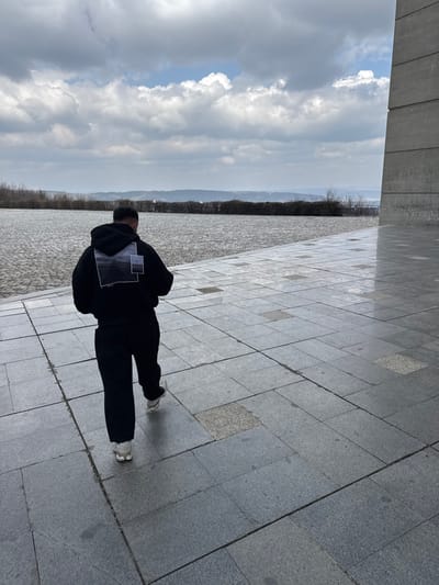 A lone male individual walks away from the camera on a large, tiled outdoor plaza at the Monument to 1300 Years of Bulgaria in Belokopitovo, Bulgaria. The individual wears a black hooded sweatshirt with a white graphic design on the back, black trousers, and light-colored athletic shoes. He is engaged with a smartphone, held in both hands, as he progresses. To the immediate right, a substantial, angular concrete structure, consistent with the monument's architecture, rises vertically. The ground is paved with varied shades of dark grey square and rectangular tiles, some appearing reflective due to moisture. The background features a low, dark green hedge separating the plaza from a distant, hazy landscape of rolling terrain under a mostly overcast sky with scattered clouds.