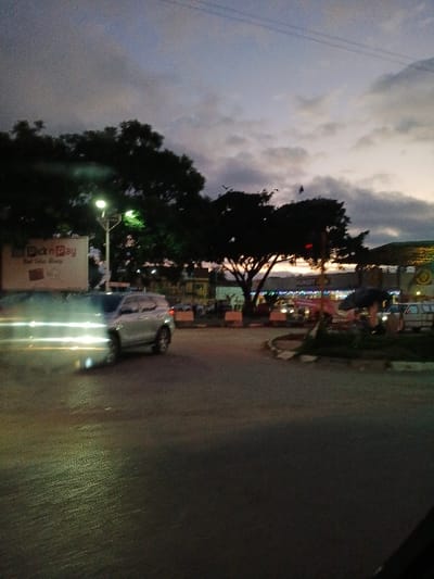 An outdoor scene at dusk. A cloudy sky hovers above trees and a commercial area with lights. A silver car with headlights on is driving on the dark street. A Pick n Pay sign is visible in the lower left corner. The image is taken from a slightly elevated perspective.