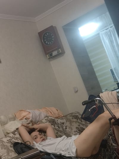 An adult female is depicted lying supine on a patterned sofa or bed within an indoor residential setting in the city of Sheptytskyi, Ukraine. She is positioned with her head towards the upper left quadrant of the frame, looking directly towards the camera with a neutral expression. Her arms are bent and raised, with her hands clasped behind her head. She wears a light-colored short-sleeved top that is slightly gathered around her waist and upper thighs. Her lower body, including her legs, extends towards the right, partially obscured by a bicycle.
The sofa or bed features a dark brown and beige paisley or floral pattern. To the left of the woman's head, on the sofa, are several items of folded fabric in peach and white, and a white stuffed animal. In the foreground, below the woman, a dark pouch or bag is visible on the sofa, alongside a dark rectangular object.
The background wall is a plain light beige. A wooden wall clock with a dark face and Roman numerals (XII, III, VI, IX) is mounted on the upper left wall, featuring a lighter, slatted section below the clock face. White crown molding is visible at the junction of the wall and ceiling.
To the right, a brightly illuminated rectangular opening, possibly a window or doorway, reveals horizontal blinds and sheer white patterned curtains. The intensity of the light source is significant. A portion of a bicycle, including handlebars with black grips and chrome levers, extends from the middle to the lower right of the frame, with a dark blue fabric draped over its frame. A small, light-colored object is affixed to the wall below the window. The overall scene suggests a lived-in private dwelling.
