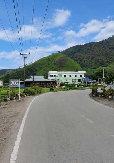 The image shows a road curving through a landscape dominated by lush green hills under a partly cloudy blue sky. On the left side of the road, there is a sign with the letters "AG" in large white block letters. Behind the sign, there are telephone poles with numerous wires stretching across the frame. Further back, a low-lying building with a corrugated metal roof is visible, and beyond that, a larger, multi-story building with a white upper section and a light green lower section. This building appears to be modern with many windows. The surrounding environment is verdant, with trees and foliage visible on the hillsides and closer to the buildings. The road is paved and has white lane markings. There are some potted plants and what appear to be stones or small rocks along the side of the road near the buildings. The overall scene suggests a rural or semi-rural setting within a mountainous region.