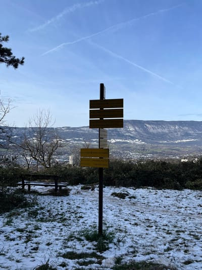 The image captures an elevated viewpoint overlooking the city of Chambéry, France, specifically in the vicinity of the Belvédère Pariset, as indicated by a directional sign pointing to "Belvédère Pariset 0.3km 6min." The scene is set during winter or early spring, with patches of snow covering the ground, interspersed with dry grass and exposed soil.
At the center, a dark brown metal pole serves as a signpost, displaying multiple yellow rectangular directional signs. These signs, written in French, point to various local destinations with associated distances and estimated walking times, including "Mont Calamin," "Le Reposoir," "Chambéry - Pont des Amours," "Sainte Claire - Curial," "Chambéry - Parc Pajots," "Belvédère Pariset," and "Belvédère Roques."
To the left of the signpost, a dark wooden park bench is visible, partially dusted with snow. The mid-ground features a scattering of bare deciduous trees and dense green evergreen shrubs. In the background, a valley unfolds, revealing the urban landscape of Chambéry with indistinct buildings, all framed by snow-dusted mountains. The sky is clear and bright blue, marked by several prominent white aircraft contrails, notably forming an "X" pattern in the upper portion of the frame. No individuals, animals, or active interactions are present in the image. The lighting suggests clear daytime conditions.