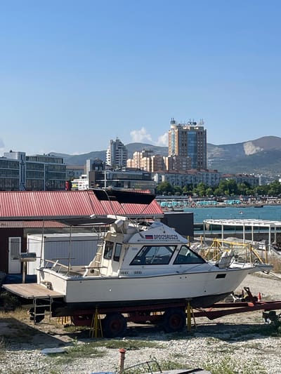 The image depicts a boat on a trailer, parked on a gravelly area in front of a building with a red corrugated metal roof. In the background, a city skyline is visible with several tall buildings, including a prominent multi-story tower with logos at the top. Beyond the city, mountains rise in the distance. A body of water, likely the sea, is visible with beach umbrellas and people on the shore. The sky is clear and blue. The boat is white, with a cabin, and has a registration number "OP0125RUS25" and a Russian flag decal on its side. The trailer appears to be red and has two wheels. The environment suggests a sunny day in a coastal city.