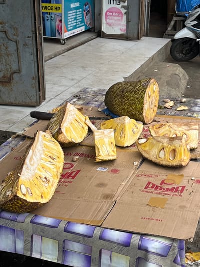 In Gamponguleegle, Indonesia, a cut jackfruit is displayed on a table covered with cardboard boxes and what appears to be decorative wrapping paper. The jackfruit is cut into several large segments, revealing its bright yellow flesh and numerous seeds. A dark-handled knife rests on one of the cardboard pieces near the fruit. In the background, a motorcycle is partially visible, suggesting a street-side market or roadside stall setting. The scene is outdoors, with a building facade in the far background that includes a sign for "SERVICE HP" (phone service) and a mobile payment advertisement, indicating a commercial area.