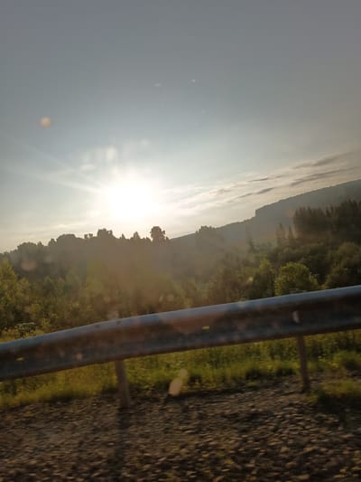 A wide-angle, slightly tilted outdoor shot captures a sun-drenched landscape, likely taken from a moving vehicle. The sun, a bright orb with visible rays, dominates the upper left portion of the frame, casting a warm, hazy glow over the scene. A dense forest covers rolling hills in the mid-ground, with trees of varying heights and shades of green silhouetted against the bright sky. In the foreground, a weathered metal guardrail runs horizontally across the image, its surface marked with rust and wear. The ground beneath the guardrail appears to be a mix of gravel and sparse vegetation. The overall impression is of a rural, possibly mountainous, environment bathed in the soft light of dawn or dusk. There are no people or animals visible in the frame. The image does not contain any information that specifically places it within the city of Богородск, Russia.