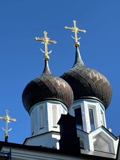 The image shows a close-up of the domes and crosses of an Orthodox church against a clear blue sky. Two large, dark, onion-shaped domes with golden crosses on top are prominent in the center of the frame. The domes appear to be made of metal or tile and are supported by white structures with light blue trim. A smaller golden cross with a crescent moon at its base is visible on a lower section of the building to the left. The architectural style suggests a Russian Orthodox church. The overall scene is bright and sunny, indicating daylight. There are no people or discernible actions taking place. The context provided of St. Ozerki, Russia, suggests this is a religious building within that location.