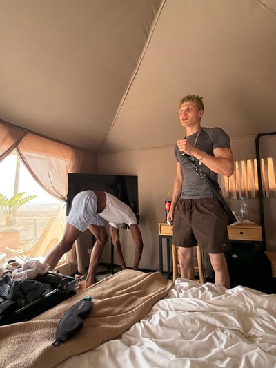 In a tent accommodation in Agafay, Morocco, a young man with blond curly hair stands looking to his right. He is wearing a grey t-shirt and brown shorts, with a black strap across his chest. To his left, another person, whose face is obscured, is bent over in a downward-facing dog yoga pose. The room is furnished with a television, a bedside table with drinks, and a bed with rumpled white sheets and a tan blanket. An open suitcase lies on the blanket, with clothes spilling out. A black eye mask rests on the blanket in the foreground. Outside the tent, a desert landscape with sparse vegetation and a lounge chair is visible through a sheer curtain.
