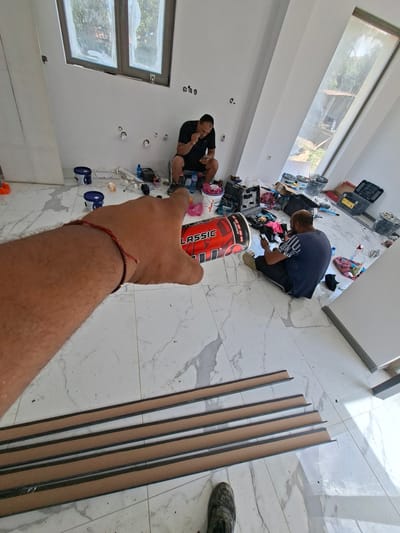The image depicts an interior scene, possibly a room under renovation, in Dolna Malina, Bulgaria. Two men are present. One man, wearing a black t-shirt and dark shorts, is squatting near a window, holding a mobile phone and what appears to be food, engaged in eating or examining the phone. The other man, wearing a blue t-shirt and dark pants, is also sitting on the floor, looking at his phone. The room features white walls and a marbled floor. Various construction materials and tools are scattered around, including paint buckets, toolboxes, pipes, and what seem to be long, narrow strips of material laid out on the floor. A prominent object in the foreground is a large, red and black cylindrical item, possibly a container or a piece of equipment, with the word "CLASSIC" visible on it. The overall impression is one of a work-in-progress environment, with the individuals taking a break from their tasks.