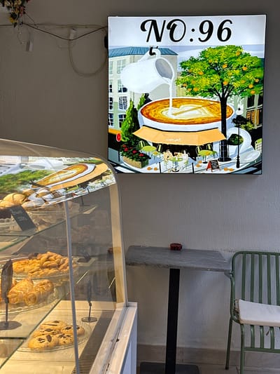 The image captures an indoor scene in Datça, Turkey, featuring a bakery or cafe. A brightly lit sign or artwork is on the wall, displaying a stylized image of a cafe scene with the number "No.96". The scene includes a painting of a coffee cup and pitcher. Below the sign is a glass display case containing various baked goods, including pastries, cookies, and other treats. A small sign is visible in front of one of the trays of baked goods. A metal table and a green metal chair are also visible. Wires run from the sign down the wall. Flowers are displayed near the bottom left of the frame. The reflection of the sign on the display case.