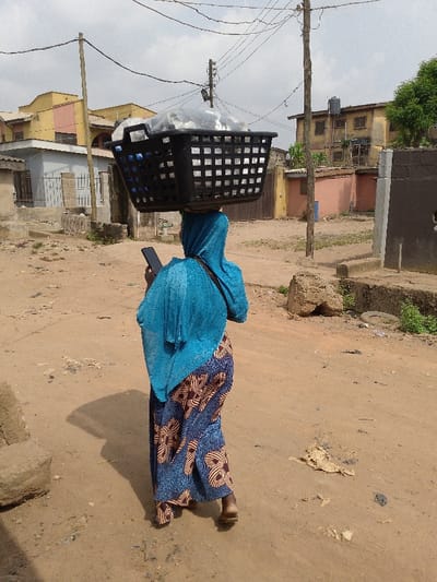 A solitary individual is observed from behind, walking on an unpaved, reddish-brown dirt road in Ijaiye, Nigeria. The person wears a teal head covering, a long patterned dress with blue, brown, and white designs, and light-colored sandals. A large, black, rectangular woven plastic basket, containing multiple items encased in clear plastic bags, is balanced on their head. A black smartphone is held in their right hand. A dark strap is visible across their right shoulder. The background features residential buildings of various states of repair; a yellow and pink multi-story structure is visible to the left, and a weathered tan two-story building with a rooftop water tank stands to the right. Two wooden utility poles with a dense network of overhead wires traverse the scene. The ground is characterized by scattered debris, including rocks, a deflated tire, and fragments of cardboard. Shadows indicate daylight conditions.