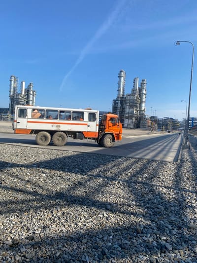 An outdoor, mid-range, slightly elevated shot captures a sunny, clear day with a vast blue sky at the Yukhta oil refinery in Russia. In the foreground, a wide, grey road paved with small, dark grey stones runs from the left to the right of the frame. A shadow cast by an unseen object stretches across this area.

A large, six-wheeled bus, painted white with an orange stripe and orange accents on the front and rear, is captured in motion, moving from left to right across the frame. The bus's windows reveal several individuals inside, some wearing orange vests, indicating they are likely workers. The bus has the markings "RENCON SF" and "+47" on its side.

In the mid-ground, the industrial complex of the Yukhta oil refinery dominates the landscape. It features numerous tall, metallic structures, including distillation towers and intricate pipe networks, all under the bright blue sky. A tall lamppost with a security camera is visible on the right side of the road, facing towards the refinery. Further in the background, more industrial structures and a clear horizon line are visible.