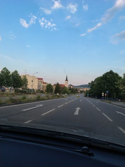 The image shows a view from a vehicle looking out at a road and its surroundings in Strakonice, Czechia. The perspective suggests the photo was taken from the passenger seat, looking forward and slightly to the left. The road, with white lane markings and arrows, curves into the distance, bordered by a grassy median and trees.
On the left side of the road, several buildings are visible, including what appears to be a church with a steeple. The architecture suggests a European setting, with tiled roofs and light-colored facades. Trees, some with dense green foliage, line the street and are interspersed with buildings. The sky is blue with scattered white clouds, indicating fair weather. A faint contrail from an airplane is visible in the upper left portion of the sky.
The time of day appears to be late afternoon or early evening, judging by the soft light and the sky's coloration. The overall scene is peaceful and everyday, depicting a typical roadside view in a town or city. There is no discernible activity or event taking place, and no people are clearly visible.
The image is slightly tilted, with the horizon line running diagonally. The foreground is dominated by the interior of the vehicle, with a dark blue surface and a portion of the windshield wiper visible. No text is clearly legible in the image.