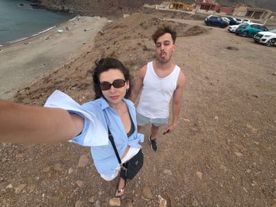 In Níjar, Spain, a young woman in a light blue button-down shirt and white shorts, wearing sunglasses and flip-flops, takes a selfie. She is accompanied by a young man in a white tank top and striped shorts, who is sticking out his tongue. They are standing on a rocky, dirt path overlooking a beach with a few scattered umbrellas and people. Several cars are parked in the background near some buildings. The scene appears to be a coastal area.
