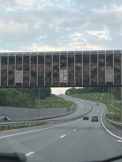 The image shows a highway in Званка, Russia. A modern pedestrian bridge with dark tinted glass and metal framing spans over the highway. The bridge appears to be a noise barrier, with ribbed metal panels visible within some of its sections, suggesting it's designed to muffle traffic sounds.

The highway itself is grey asphalt with clear white lane markings, curving gently to the right. Several cars are visible on the highway, driving in the same direction. A few streetlights are positioned along the side of the road.

To the left of the highway, there is lush green grass and trees, indicating a rural or semi-rural setting. In the background, beyond the trees, more of the highway and surrounding landscape can be seen.

The sky is partly cloudy, with patches of bright sky visible through lighter clouds, suggesting it might be late afternoon or early evening. There is no visible text in the image. The scene appears to be a typical day on a major road, with the pedestrian bridge serving as an interesting architectural element against the natural landscape.