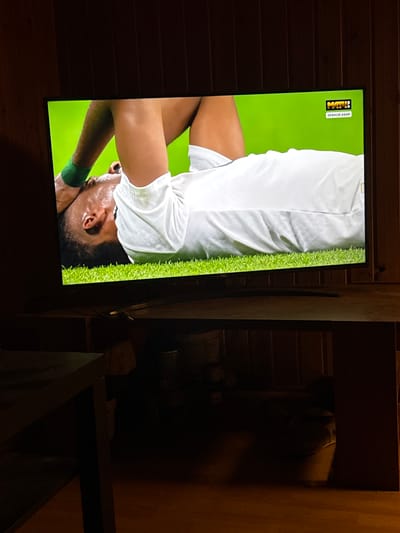 In the city of Bykovo, Russia, a television screen displays a sports broadcast. The screen shows a close-up of a Black male athlete lying on his back on green grass, appearing to be in distress or exhausted. He is wearing a white jersey and a green wristband. A watermark in the upper right corner of the screen reads "МАТЧ!" and "ПРЯМОЙ ЭФИР", indicating it's a live broadcast on the Match TV channel. The television is placed on a dark wooden table. The background is a dark, paneled wall. In the foreground, to the left, a dark wooden table or piece of furniture is partially visible. The overall lighting is dim, with the primary illumination coming from the television screen itself.