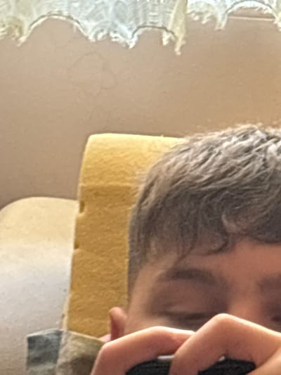 A close-up, oblique angle image shows a person's head and hand. The person has gray hair and is resting their head on a yellow, porous object. The hand is holding a black object, possibly a phone. The background is a blurry beige wall with a decorative white edge. No information in the picture indicates that this picture was taken in Damianitsa, Bulgaria.