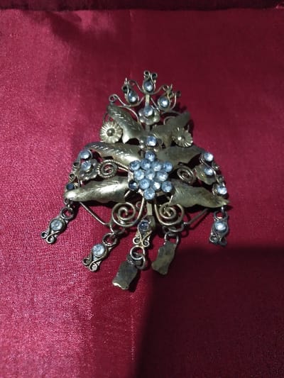 The image displays an ornate, antique-style metal brooch or pendant centered on a deep red fabric background, exhibiting visible folds and creases. The object features a central cluster of approximately nine light-colored, faceted stones, likely rhinestones, arranged circularly. This central element is surrounded by stylized metallic leaves and scrollwork, which are also set with individual light-colored faceted stones. The metal appears to be an aged brass or bronze, showing some patina. Several smaller, chain-like elements, each containing an individual faceted stone, and two plain, rectangular metallic pieces dangle from the lower section of the main structure. The lighting is diffuse, casting subtle shadows. No individuals, actions, interactions, or specific environmental features are discernible. The image does not contain visual identifiers for the city of Domažlice, Czechia.