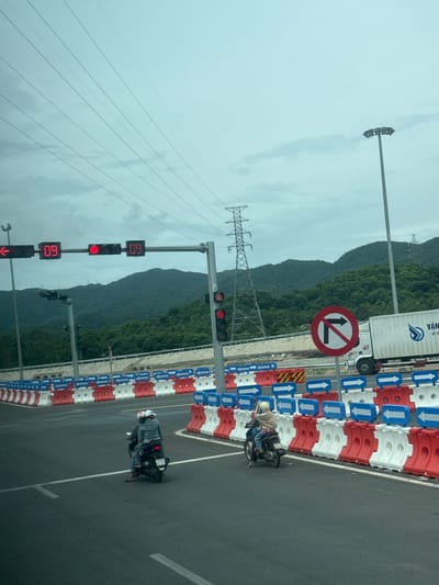 The image depicts a traffic scene in Hoa Xuan, Vietnam, likely near a construction zone or a point of altered traffic flow. In the foreground, two individuals are riding on a single scooter. The driver and passenger are both wearing casual clothing, with the passenger wearing a light-colored helmet and the driver wearing a darker helmet. The scooter has a license plate that appears to be from Vietnam. The road itself is paved asphalt, with white lane markings.
The immediate surroundings are marked by a series of traffic barriers. These barriers are a combination of red and white plastic units, some of which have blue directional arrows indicating traffic flow. These barriers create a channel or separation on the road.
In the mid-ground, there are several traffic signals. One set displays red lights and digital readouts showing "09". Another traffic signal has a red light illuminated. A prominent "no right turn" sign is visible, which is circular with a red border and a red slash over a black arrow pointing to the right.
A large white truck, possibly a container truck, is positioned to the right of the "no right turn" sign. The truck has lettering on its side, partially visible. In the background, a power transmission tower is visible, and beyond that, a densely forested mountain range under an overcast sky. The overall impression is of a functional, albeit potentially temporary, traffic management system in an outdoor, semi-urban or suburban environment.