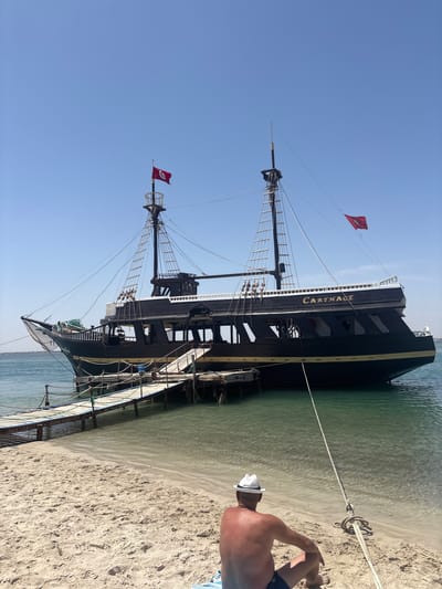 A wooden pirate-style ship, named "CARTHAGE" in gold lettering, is docked at a pier on a clear, sunny day. The ship is dark-colored with a white upper deck and features two masts with rigging. A red flag with a crescent moon and star is visible on one of the masts. A man wearing a white hat and dark swimming shorts is sitting on a sandy beach, facing away from the camera, looking out at the water. A wooden pier extends from the beach to the ship. The water is calm and clear, with the sandy seabed visible. The sky is a bright, cloudless blue. The scene appears to be in a coastal area, likely a tourist destination.