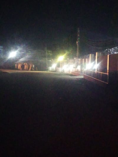 The image depicts an outdoor scene at night in Baccure Kofare, Nigeria. The foreground is dark, indicating a natural ground surface like grass or dirt. In the middle ground, a dimly lit area reveals several structures, possibly stalls or temporary shelters, with people visible in silhouette against the illuminated interiors. Bright lights, likely from lamps or floodlights, illuminate these structures and create a hazy glow in the surrounding atmosphere, suggesting a gathering or event. A fence or wall, featuring vertical posts and horizontal railings, runs along the right side of the middle ground, with more bright lights positioned behind it. A utility pole with some decorative lights, possibly for an event, is visible in the background between the structures and the fence. The overall impression is of a community gathering or market taking place after dark.
