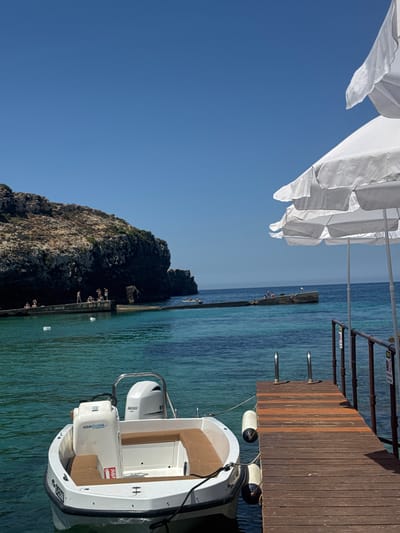 The image depicts a scenic view of a tranquil bay in Mellieħa, Malta, under a clear blue sky. In the foreground, a white motorboat with brown seating is moored to a wooden jetty. The boat features an outboard motor labeled "200" and has "AQUAMARINE" written on its console. The jetty, made of weathered wooden planks, has a metal railing and two white fenders attached to it.
Beyond the boat and jetty, the turquoise water of the bay is visible, leading towards a rocky coastline. Several people are scattered along a concrete breakwater extending into the water, some sitting and others appearing to be preparing to swim or have just emerged from the water. In the distance, a larger rock formation rises from the sea, partially covered in greenery. The overall atmosphere is one of serene summer leisure, characteristic of coastal Malta.