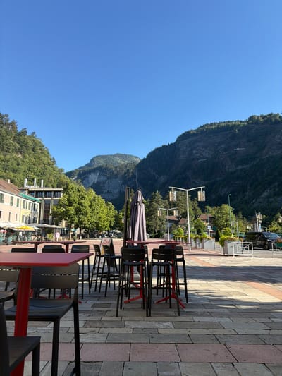 This media file displays an outdoor scene featuring a mountain range in the background and a patio area with tables and chairs in the foreground. The scene is bathed in bright sunlight, indicating it is daytime, likely late morning or early afternoon given the length of the shadows. The sky is a clear, vibrant blue, suggesting fair weather.
The mountain slopes are covered in lush green trees, with rocky outcrops visible. Below the mountains, there are buildings with light-colored facades and windows. A street with parked cars and some pedestrians is visible further down.
In the foreground, several tables and chairs are arranged on a stone-tiled patio. The furniture appears to be modern, with a mix of red and black accents. Some of the tables are set up for dining, with umbrellas partially covering some of them. The overall impression is that of a cafe or restaurant seating area.
There is some text visible on a sign placed between tables, but it is too blurry to be legible. The scene conveys a relaxed and pleasant atmosphere, typical of a sunny day in a scenic, possibly European, town. The location context of Cluses, France, suggests a mountainous region known for its natural beauty.