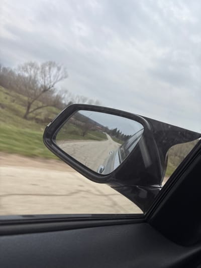 The image depicts a view from the interior of a vehicle traveling on an unpaved road near Suhache, Bulgaria, under an overcast sky. The foreground shows the upper section of a dark vehicle door panel and window frame. To the left, a grassy hillside with multiple bare deciduous trees slopes upward. The ground immediately adjacent to the vehicle appears to be a light-colored dirt or gravel road with visible surface variations. The main subject is a black exterior side mirror positioned on the right side of the frame. The mirror's glass surface exhibits several linear scratches. Reflected within the mirror is a continuation of the unpaved road, receding into the distance. Alongside the reflected road, various trees and vegetation are visible, and in the far distance of the reflection, small structures with reddish roofs are discernible among the trees. No individuals are visible. The scene suggests a daytime setting during late winter or early spring.