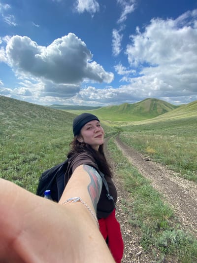 An individual is taking a selfie while walking on a dirt path through a grassy, hilly landscape under a partly cloudy blue sky. The individual is wearing a black beanie, a brown long-sleeved shirt, and a red garment visible under the shirt. A black backpack is worn by the individual. A tattoo is visible on the person's left arm, and a silver bracelet is on their wrist. The landscape features rolling green hills covered in grass, with a distinct path winding through them. The location is described as Andreevka, Russia.
