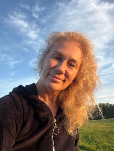 This is an outdoor selfie featuring a smiling woman with blonde, curly hair. She is looking directly at the camera with a gentle smile, her face and hair illuminated by a warm, golden light, suggesting late afternoon or early evening. She is wearing a black, possibly velvet, zippered top, with a silver zipper and ring-shaped pull visible on her right shoulder.

The image is noticeably tilted. The background on the left shows a clear blue sky with scattered white, wispy clouds. On the right, behind the woman, lies a lush green field extending into the distance, with trees visible along the horizon. The scene conveys a relaxed and pleasant atmosphere in a natural setting. There is no visible text in the image.