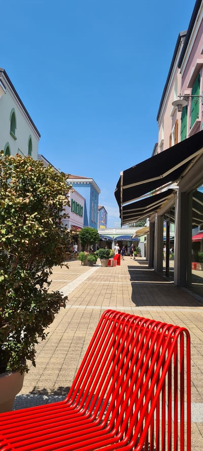 This is a vertical, eye-level medium shot of a picturesque outdoor shopping area on a sunny day. The scene is an open-air promenade lined with colorful buildings, likely boutiques or shops. On the right side of the frame, a striking bright red metal bench dominates the foreground, its curved tubular design casting sharp shadows on the light-colored tiled pathway.
The pathway is paved with square tiles and a prominent white stripe runs down its center, creating a sense of perspective as it leads into the distance. Along the left side of the path are awnings extending from the buildings, some dark and some lighter, offering shade. Beyond the awnings, buildings display a variety of pastel colors, including pink, light blue, and green. One building prominently features green shutters.
In the middle ground, several people are visible walking and browsing, suggesting a relaxed atmosphere and leisure activity. A "Tommy Hilfiger" sign is visible on one of the shops, providing a specific location cue. Small trees and bushes are strategically placed throughout the area, adding greenery to the scene. The sky is a clear, vibrant blue, indicating fair weather and likely daytime, possibly late morning or early afternoon due to the distinct shadows.
The overall impression is one of a bright, inviting commercial district designed for pleasant strolling and shopping. The composition, with the bold red bench in the foreground and the receding line of shops, draws the viewer into the scene. The clear sky and sunshine evoke a feeling of warmth and enjoyment.