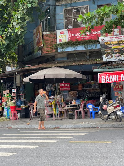 The image depicts a street scene in Tân Quang Commune, Vietnam, focusing on a roadside stall. A man, appearing to be in his late 40s or early 50s, with short, dark hair and a slim build, stands on the sidewalk, drinking from a can and smoking a cigarette. He is wearing a short-sleeved, olive-green polo shirt and dark-colored, knee-length shorts, and is barefoot. The stall is covered by a large, gray umbrella, and the facade of the building behind it is painted light blue. The stall displays various goods, including drinks, snacks, and what appear to be household items. Several plastic chairs, a mix of red and blue, are arranged around tables in front of the stall. A beige scooter is parked to the right of the scene. In the background, signs in Vietnamese indicate businesses, including one advertising "BÁNH MÌ" (bread) and another for "THƯƠNG QUANG" (Thương Quang). The street in the foreground features a pedestrian crossing. Trees with lush green foliage overhang the scene from the left.