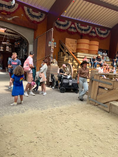The image depicts an outdoor scene with a sandy ground and a paved area, likely in an amusement park or entertainment venue, given the rustic wooden structures, decorative banners, and presence of multiple people. In the foreground, a young girl with curly hair is wearing a blue dress with a rainbow on the back and white sneakers. To her left, a man in a blue t-shirt stands near an entrance. Behind the girl, a man in a pink shirt and shorts carries a pink backpack. A woman in a grey shirt and skirt stands to the right of the man with the backpack, holding clothing.

Further into the scene, a woman in a brown shirt, light-colored pants, and a cowboy hat walks towards the right, appearing to be an employee or performer. She is near a wooden structure and a large wagon. Several people are seated on wooden bleachers in the background, some appearing to be watching an event. Two large wooden barrels are visible behind the bleachers. The overall environment suggests a themed area, possibly with a Western or American frontier motif, indicated by the decorative elements and wooden construction. The location is not specified as Collendoorn, Netherlands in the image.