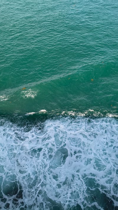 An aerial perspective shot shows the turbulent, turquoise waters of the ocean. The image is divided into two distinct sections: the upper portion consists of calmer, deeper water, characterized by subtle ripples and the presence of scattered brown organic matter. The lower half is dominated by the white froth and foam of breaking waves. These waves create intricate patterns of swirling white water against the darker, deeper ocean. There are no people or man-made objects visible in the frame. The environment depicted is exclusively natural ocean water.