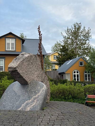The image shows a street scene in Reykjavik, Iceland. In the foreground are two stone sculptures, one blocky and textured, the other smooth and rounded, set amidst greenery. Behind the sculptures are colorful wooden buildings, typical of Icelandic architecture. The buildings have yellow and mustard-colored facades with grey roofs and white-framed windows. One building has distinctive arched windows. The street is paved with interlocking stones. The sky is overcast. No people are visible in the frame. The location is likely in central Reykjavik, known for its colorful buildings and public art.