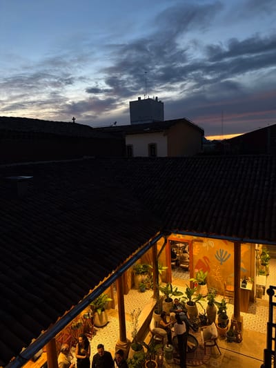 An elevated view of a courtyard gathering beneath a tiled roof, illuminated with warm light amid potted plants and patterned flooring. Adjacent, a dark, tiled rooftop leads to a building with a white structure. The sky features a gradient of colors, from dusky orange near the horizon to a pale blue speckled with clouds.