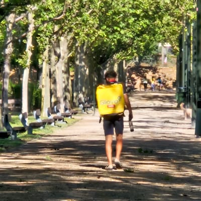 A person wearing a yellow delivery backpack walks down a tree-lined path in Zaragoza, Spain. The person is wearing dark shorts and light-colored shoes. The path is made of dirt and gravel, and is dappled with sunlight and shadows from the trees. There are benches on either side of the path. In the background, other people are walking and cycling. The trees are full of green leaves, and the sky is bright and sunny. The overall scene is one of a pleasant day in a park or urban green space.
