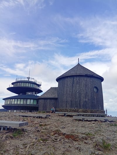 The image captures a striking view of the meteorological observatory at Sněžka, the highest peak in the Czech Republic, situated near Malá Úpa. Two distinct architectural structures dominate the scene. On the left is a modern, multi-tiered circular building with extensive glass windows and a metallic railing around its upper level, topped by an antenna. To its right stands a rustic, cylindrical wooden building with a conical shingled roof and a small circular window. Both buildings are constructed on a rocky, uneven terrain, with wooden walkways and benches in the foreground, suggesting a tourist or observation area. A few individuals are visible, one seated on a bench near the wooden structure and others further in the distance. The sky above is a pale blue, dotted with wispy white clouds. The overall atmosphere is one of a high-altitude, remote location, likely a popular spot for its panoramic views and scientific significance.