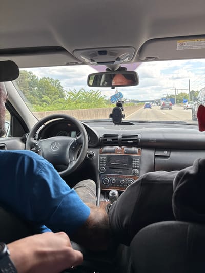 The image depicts the interior of a Mercedes-Benz vehicle in motion on a highway, likely near Zeppelinheim, Germany, given the context. The driver, a person with visible stubble, is wearing a blue shirt and appears to be driving. Their hands are on the steering wheel. In the passenger seat, another individual is visible from behind, wearing dark clothing. The dashboard features a Mercedes-Benz logo on the steering wheel, a car stereo, air vents, and a gear shift. A phone mount is attached to the windshield, and a blue car-shaped air freshener hangs from the rearview mirror. Through the windshield, the highway is visible with other vehicles and trees lining the roadside. The sky is cloudy.