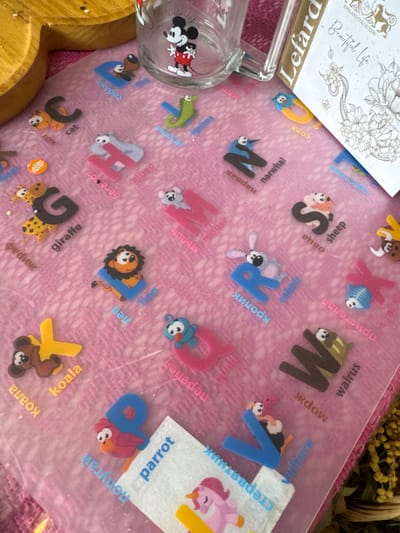 A clear glass mug featuring a red-shorted Mickey Mouse graphic is positioned on a translucent pink educational mat in the city of Matveevskoe, Russia. The mat displays the English alphabet with corresponding animal figures and names in both English and Cyrillic script. Visible elements include the letters 'B' with a dog, 'C' with a cat, 'G' with a giraffe, 'H' with a hippo, 'I' with an iguana, 'K' with a koala, 'L' with a lion, 'M' with a mouse, 'N' with a narwhal, 'P' with a parrot, 'R' with a rabbit, 'S' with a sheep, 'V' with a vulture, 'W' with a walrus, and 'X' with a fish, each accompanied by its English and Russian name. A small white label with the word "parrot" is partially visible over the 'P' section, also showing a partial unicorn graphic. To the upper right, a white booklet or card titled "Lefard ENGLAND COLLECTION Beautiful Life," featuring an ornate floral and possibly reptilian design, stands upright. A partial wooden object is visible in the upper left, and dried yellow foliage is partially visible along the right edge. The pink mat and wooden object exhibit scattered glitter particles. No individuals are present.