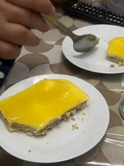 In Ksar-el-Kebir, Morocco, an indoor scene captures a close-up view of two dessert plates on a patterned surface. A human hand, positioned in the upper left, grips a metal spoon. The spoon is actively engaged with a portion of a bright yellow, glossy-topped dessert resting on a white circular plate in the mid-ground right. This dessert portion is partially consumed, revealing a lighter, crumbly base beneath the yellow layer, with some crumbs scattered on the plate. In the foreground left, a larger, rectangular slice of the identical yellow-topped dessert sits intact on another white plate, also displaying its layered structure and surrounding crumbs. The underlying surface is a brown and beige patterned tablecloth or laminate. In the blurred background, a black television remote control with multi-colored buttons is visible.