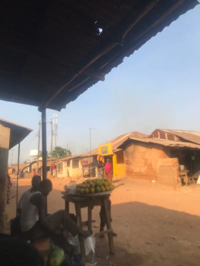 The image captures a street scene in Kwal, Nigeria, under a bright, clear sky. In the immediate foreground, a dark, corrugated metal roof, visibly worn with several holes, extends from the upper left, supported by a prominent wooden pole. Beneath this shade, two male figures are present; one, wearing a white tank top, is seated on a low surface, facing away from the camera, with his right foot resting on a white plastic bag on the ground. A wooden table is positioned centrally, holding a large, round metal tray piled high with numerous green-yellow spherical fruits, consistent with oranges.
The midground features a wide, unpaved area or dirt road. Along the left and extending into the background, a row of single-story buildings with earthen or mud-colored walls and corrugated metal roofs are visible. In the distance, a tall communication tower rises above these structures. To the right, a distinctive bright yellow building with a blue and yellow sign above its entrance stands, with a person in pink attire near its doorway. Adjacent to this yellow building, another structure with reddish-brown earthen walls and a corrugated metal roof is present, featuring a small shaded area or alcove.