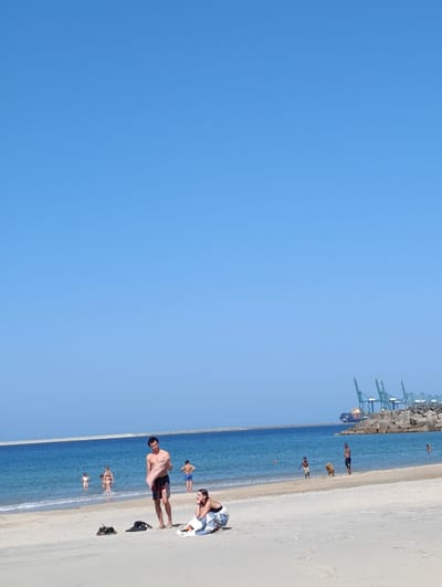 The image depicts a sunny day at the beach in Sines, Portugal, with a clear blue sky and calm ocean waters. In the foreground, a man is standing on the sand, holding a towel. Nearby, a woman is sitting on the sand, leaning back on her arms. Other beachgoers are visible in the shallow water, including children and adults. In the background, a long breakwater stretches across the ocean, and a large cargo ship and several cranes are visible at the port of Sines, with a rocky breakwater also present. The sand is light in color, and the water is a vibrant blue. Several items of clothing and accessories are scattered on the sand in the foreground.