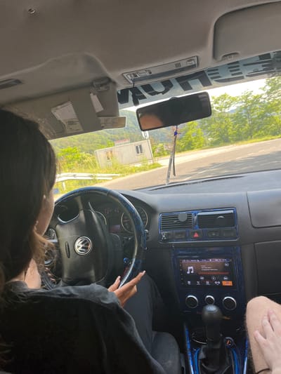 This is a first-person perspective image taken from inside a car, likely a Volkswagen, with the driver and passenger visible. The driver, a young woman with long dark hair, is seated on the left, her hands on the steering wheel. Her passenger, a person with fair skin and visible toenails, is seated on the right, with their hand resting on their leg.
The car's interior features a black dashboard with blue accents and a multimedia system displaying a song titled "Runaway" by Kanye West. The steering wheel is black with a blue patterned cover and a Volkswagen logo. Outside the car, a scenic view of rolling green hills and trees is visible, with a white portable building or structure on the right side of the road. The road appears to be paved and winding. The image was taken during daylight, with bright sunlight filtering into the car.
While the precise location in Leshtak, Bulgaria is not explicitly indicated in the image itself, the context of the question implies this setting. The image captures a moment of travel, with the passengers enjoying the scenery and music.