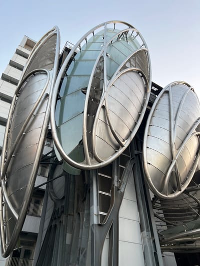 A low-angle shot reveals a building's exterior adorned with oval-shaped, metallic structures. These structures appear to be partially enclosed with glass panels. The building is modern, with visible rectangular windows on its side. Metal beams and supports intricately connect the oval features, suggesting a complex architectural design. The sky is a light, pale blue.